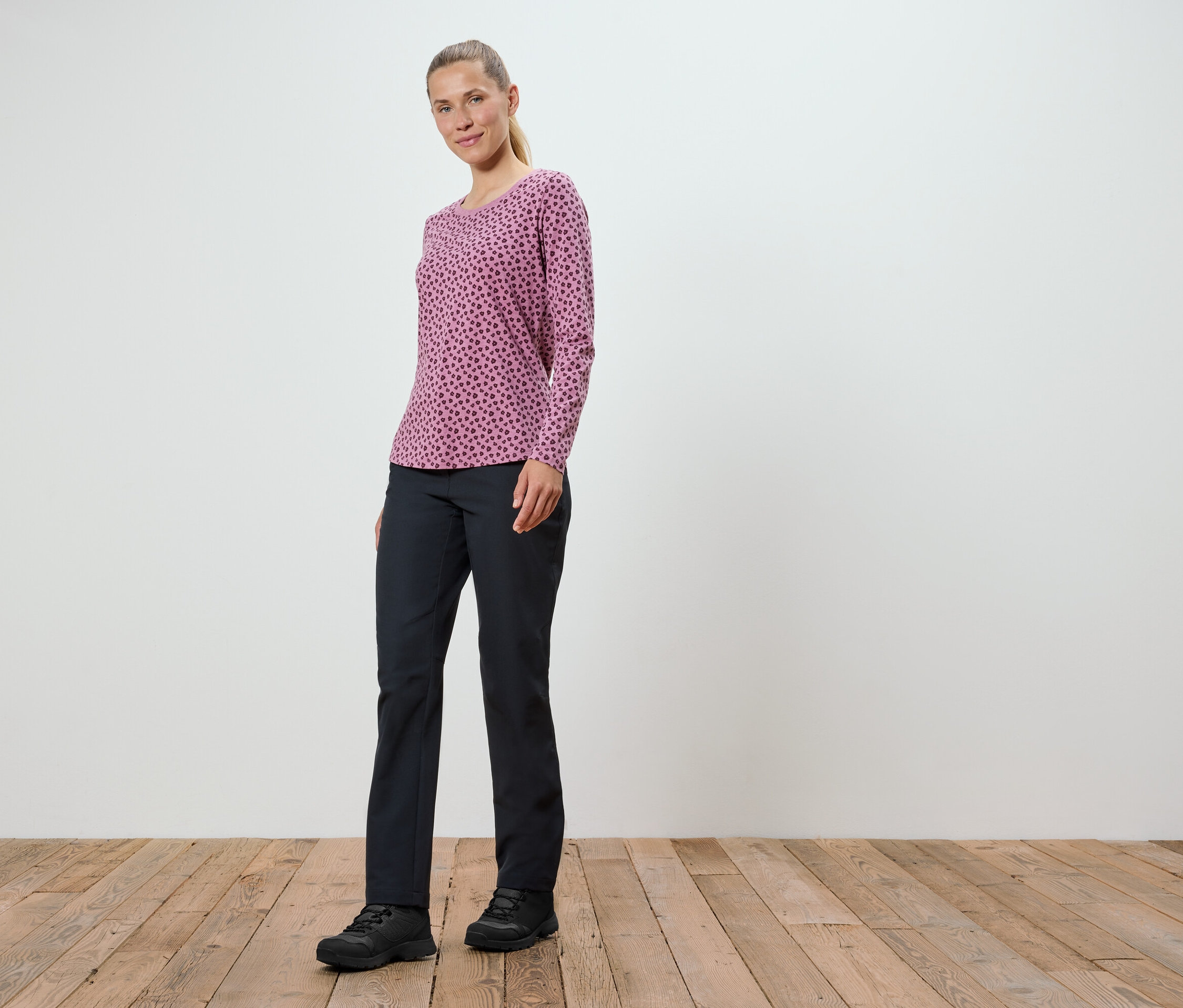 Eine Frau in einem rosa Langarmshirt und schwarzen Hosen steht auf einem Holzboden.