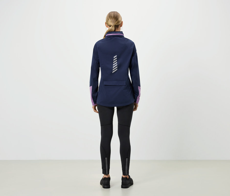 Rückansicht einer blonden Frau in blauer Jacke mit lila Ärmeln, schwarzen Leggings und schwarzen Schuhen.