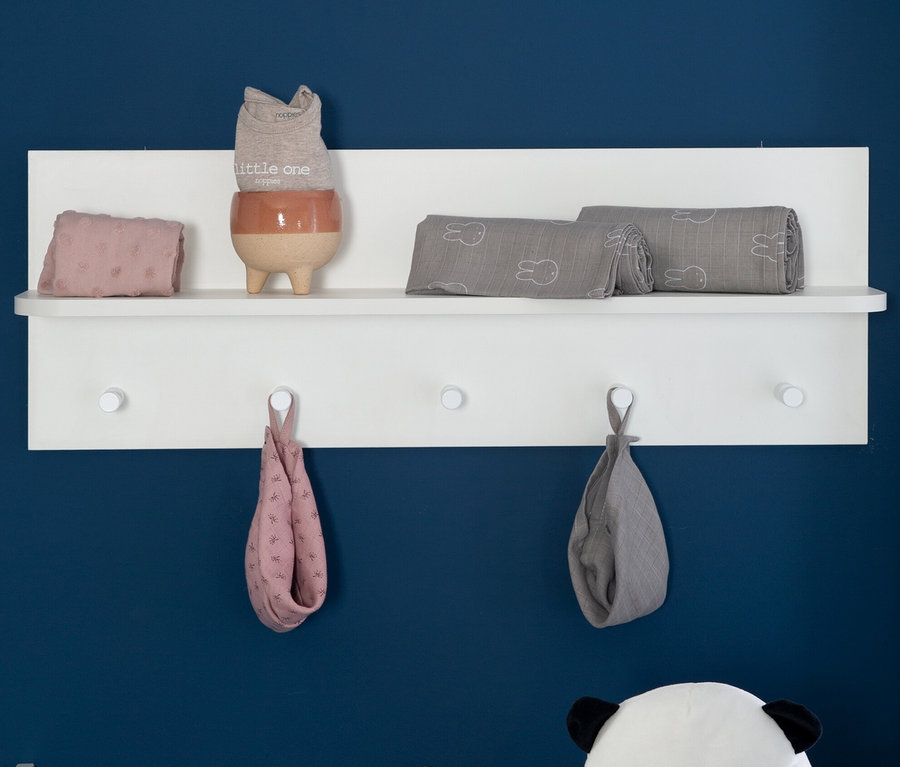 Auf einem weißen Regal liegen gefaltete Kinderstoffe und Kleidungsstücke, darunter hängen zwei Tücher an Haken.