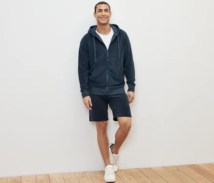 Mann steht und trägt eine blaue Frottee-Kapuzenjacke, T-Shirt und Shorts.