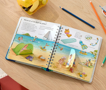 Aufgeschlagenes Buch »Wieso? Weshalb? Warum? junior: Wir fahren in den Urlaub« zeigt Illustrationen von einem Strand mit Menschen und Strandutensilien.