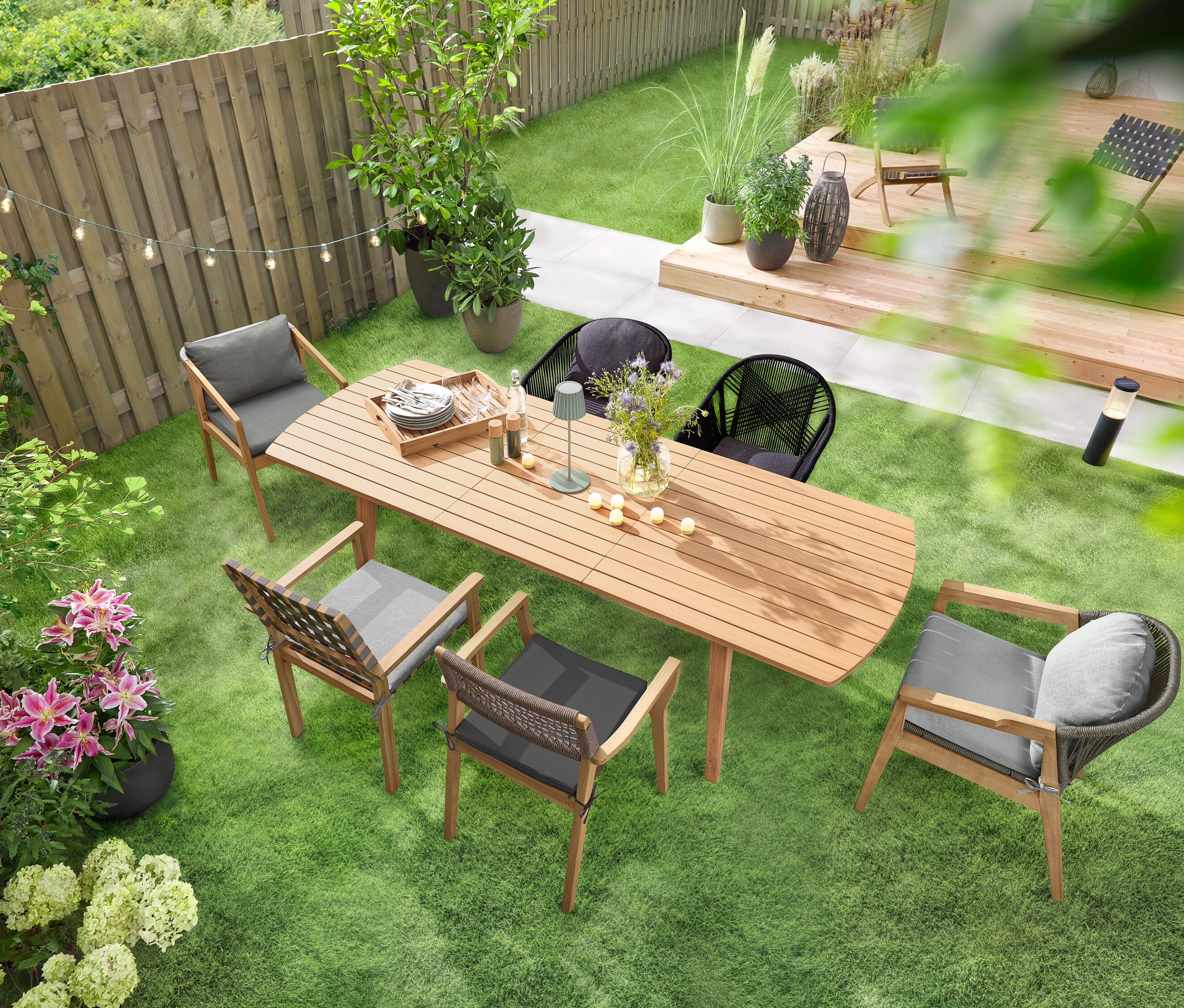 Ein ausziehbarer Gartentisch mit abgerundeten Fronten steht auf einer grünen Rasenfläche, umgeben von mehreren Gartenstühlen. Eine Solar-Lichterkette »Glühbirnen« hängt über dem Tisch, und eine LED-Akku-Outdoor-Tischleuchte, salbei, steht darauf. Mehrere 6 LED-Outdoor-Akku-Teelichter sind auf dem Tisch verteilt.