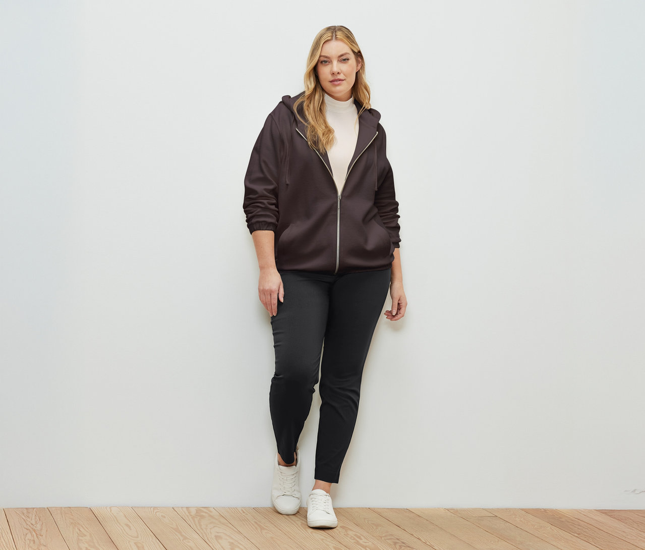 Eine Frau steht vor einer Wand und trägt eine dunkelbraune Sweatjacke.