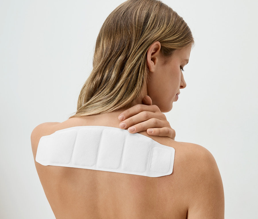 Frau mit blonden Haaren hat ein weißes Wärmepflaster auf dem Rücken und berührt ihren Nacken.