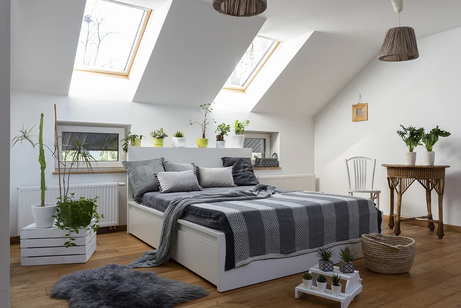 Schlafzimmer mit weißem Bett, grauen Kissen und Decke, sowie Pflanzen und Holzboden.