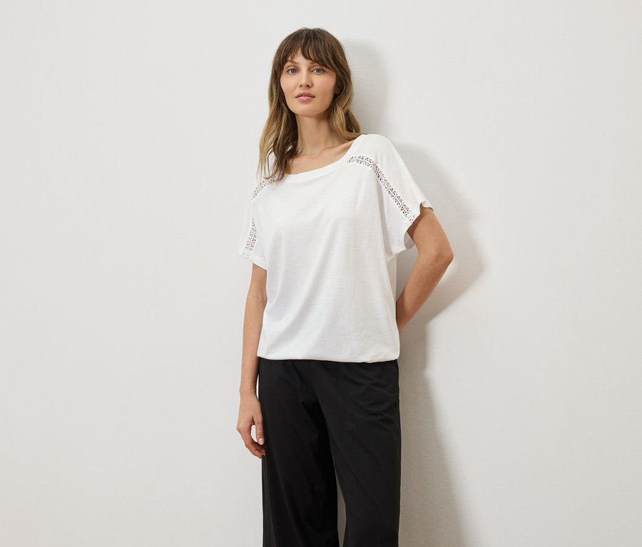 Frau trägt ein weißes T-Shirt mit Lochstickerei an den Ärmeln und eine schwarze Hose.