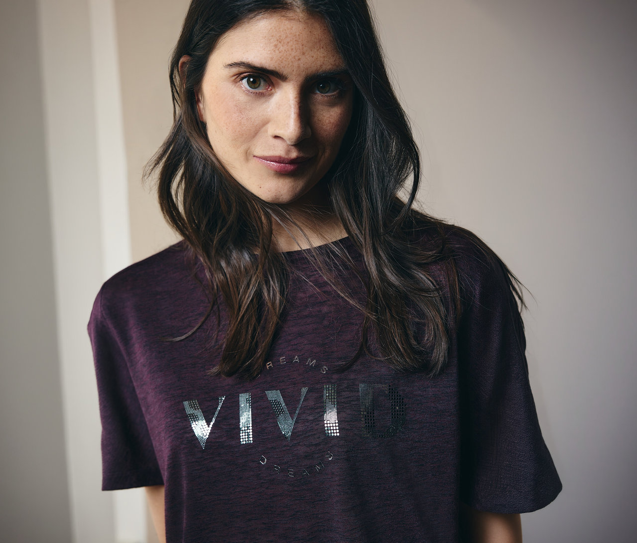 Frau mit langen braunen Haaren trägt ein kastanienbraunes T-Shirt mit der Aufschrift "Vivid Dreams".