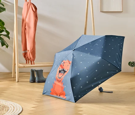 Ein blauer Regenschirm mit Dinosaurier-Motiv steht auf einem Holzboden, daneben eine Garderobe mit Regenkleidung und Gummistiefeln.