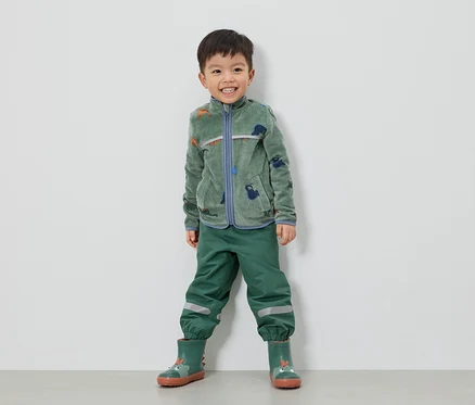 Ein lächelnder Junge steht in grüner Fleecejacke mit Dinosauriern, grüner Hose und grünen Gummistiefeln vor einer weißen Wand.