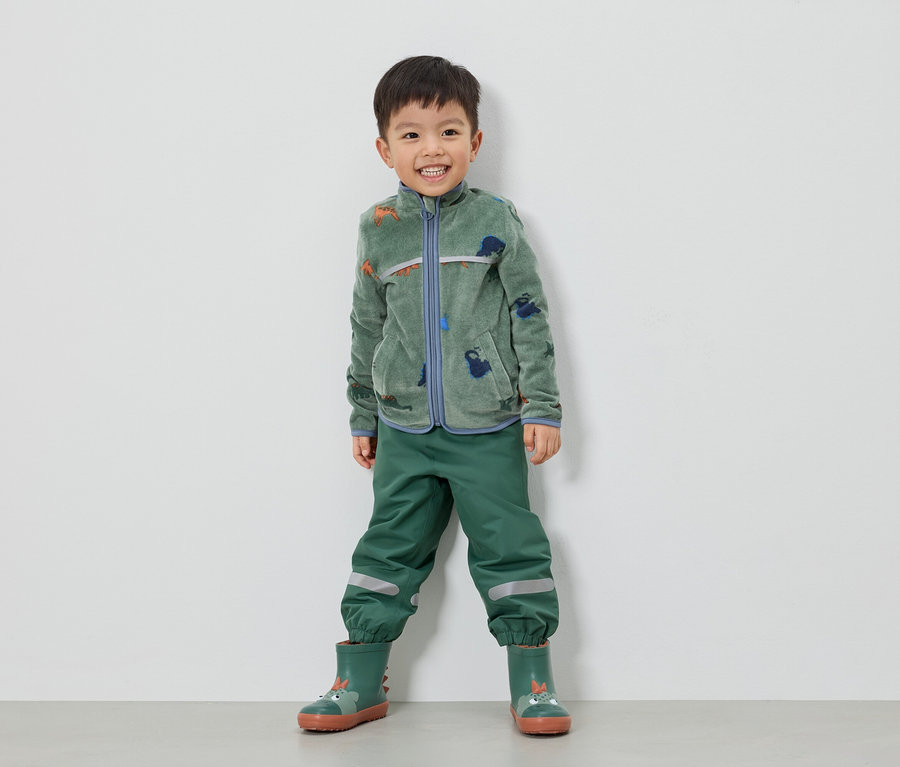Ein lächelnder Junge steht in grüner Fleecejacke mit Dinosauriern, grüner Hose und grünen Gummistiefeln vor einer weißen Wand.