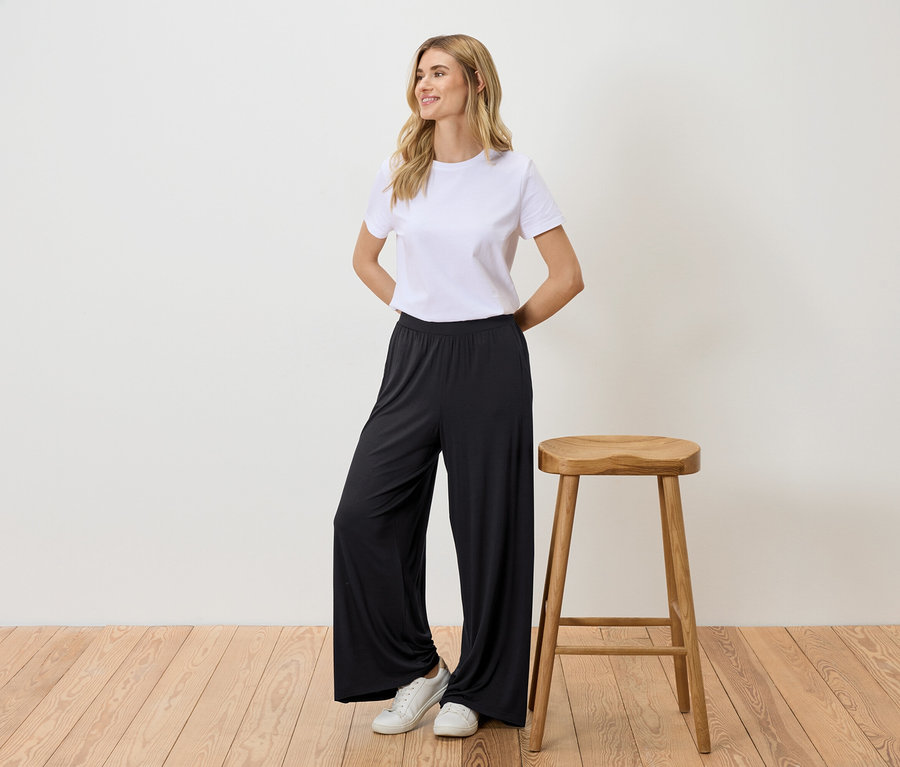 Frau steht neben einem Hocker, trägt ein weißes T-Shirt, eine schwarze Hose und weiße Turnschuhe.