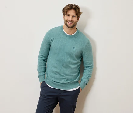 Mann mit hellbraunem Haar und Bart, der einen hellblauen Pullover und eine marineblaue Hose trägt und lächelt.