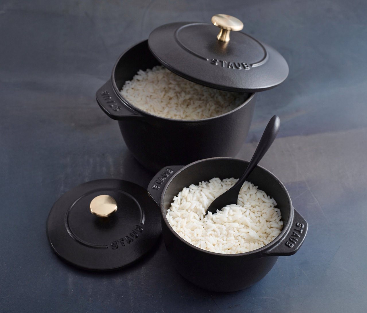Zwei schwarze, runde STAUB Reis-Cocotten (16 cm) stehen auf einer grauen Oberfläche, eine davon mit einem schwarzen Löffel.