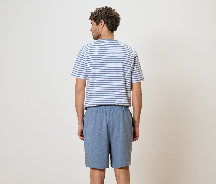 Rückansicht eines Mannes mit lockigem Haar, der ein blau-weiß gestreiftes T-Shirt und blaue Shorts vor einem cremefarbenen Hintergrund trägt.