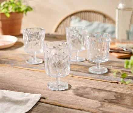 Vier klare, facettierte Gläser auf einem Holztisch, zusammen mit einer Wasserflasche, einem Teller und Topfpflanzen.