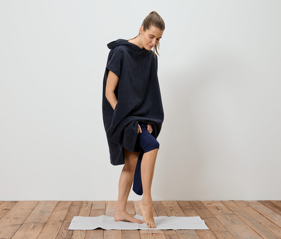 Frau im marineblauen Badeponcho, der bis zu den Oberschenkeln reicht, steht barfuß auf einer Yogamatte.