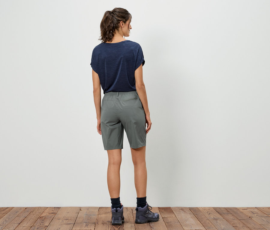 Rückansicht einer Frau mit dunklem T-Shirt, grauen Shorts und Wanderschuhen vor einer weißen Wand.