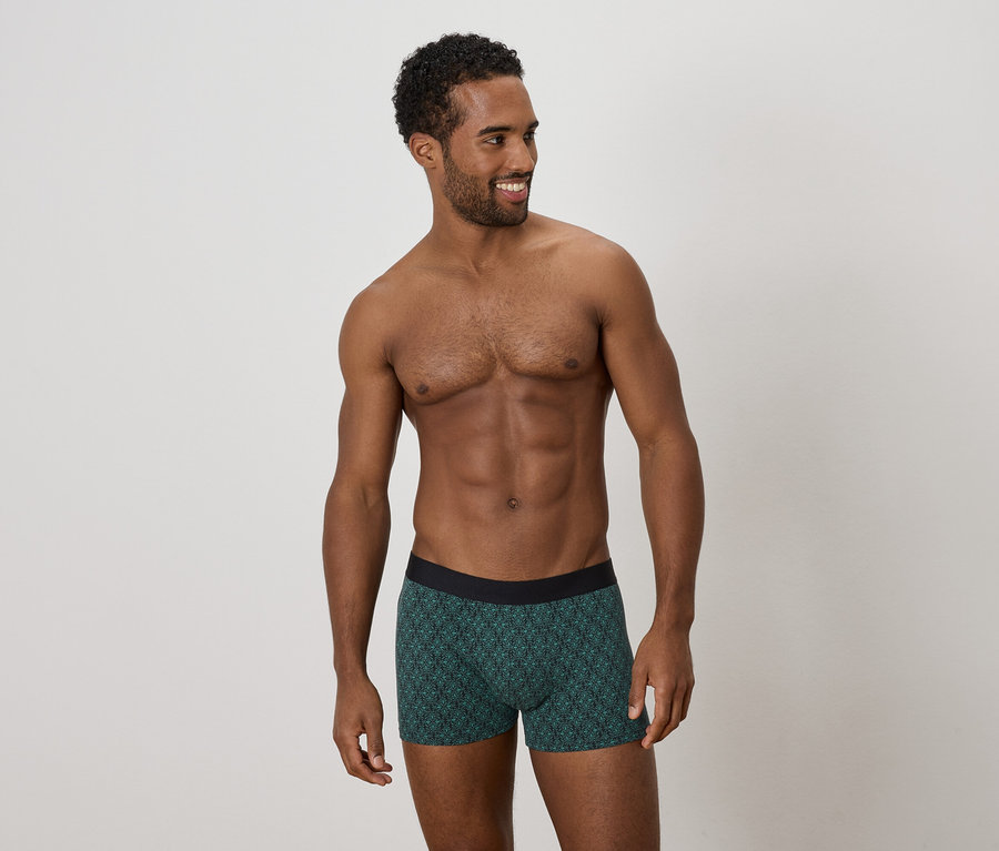 Mann mit freiem Oberkörper, der grüne Boxershorts mit Muster trägt und zur Seite schaut.