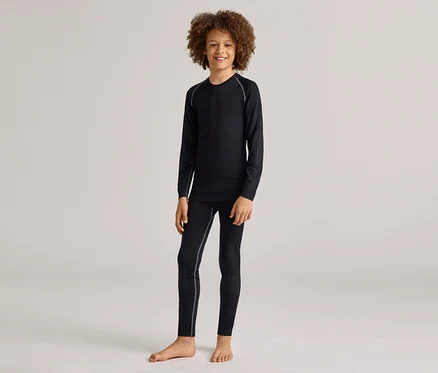 Ein lächelnder kleiner Junge mit lockigem Haar steht barfuß und trägt ein schwarzes Langarmshirt und passende schwarze Leggings mit hellgrauen Nähten.