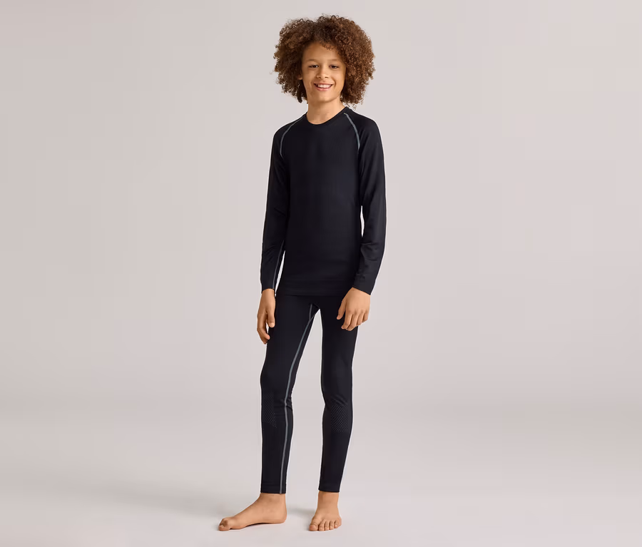 Ein lächelnder kleiner Junge mit lockigem Haar steht barfuß und trägt ein schwarzes Langarmshirt und passende schwarze Leggings mit hellgrauen Nähten.