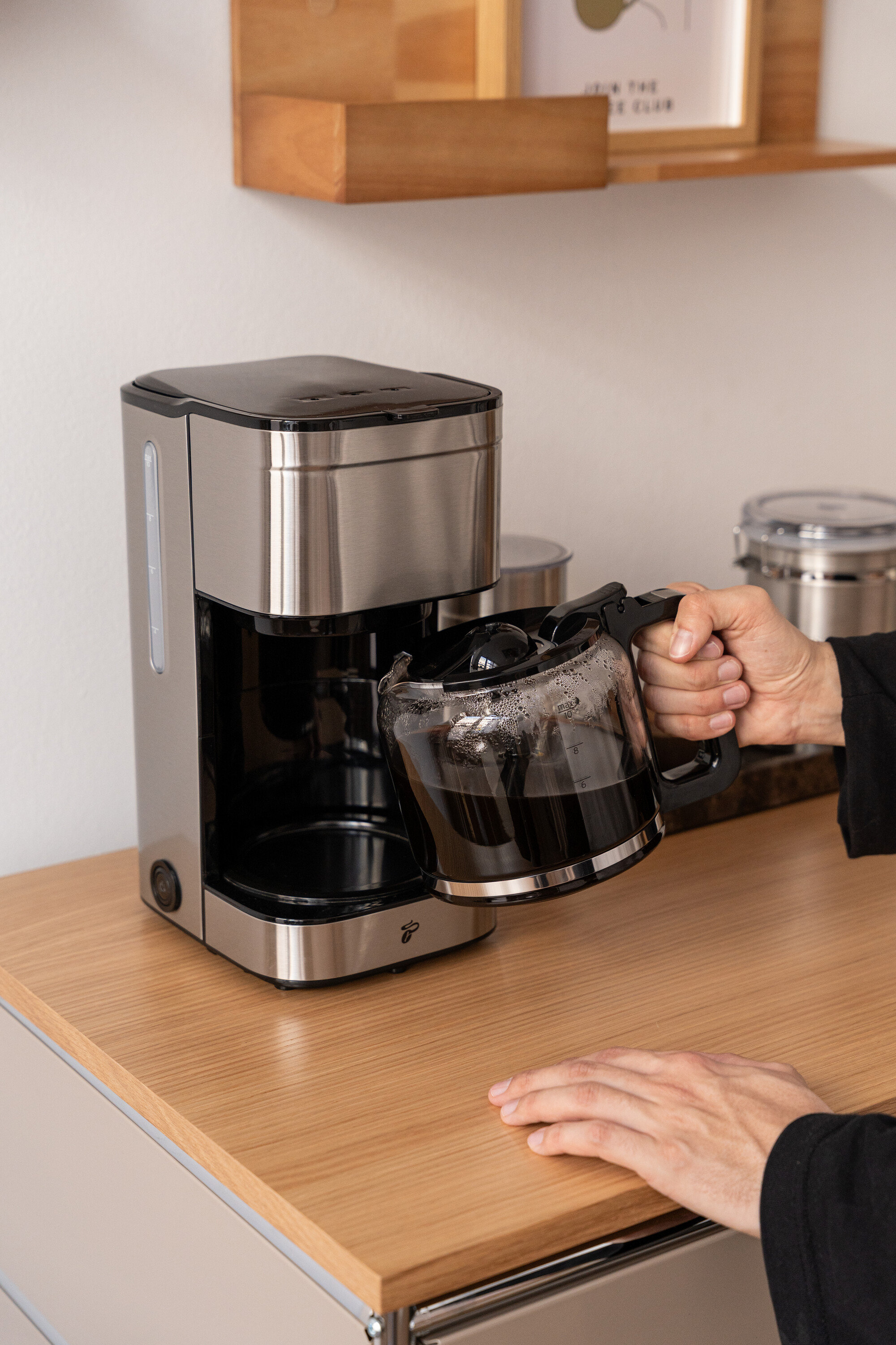 Eine Person hält eine Glaskaraffe mit Kaffee neben einer silberfarbenen Kaffeemaschine auf einer hellbraunen Arbeitsfläche.