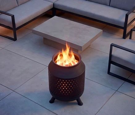 Eine dunkle, zylindrische Feuerschale mit perforiertem Design und hellen Flammen steht auf einer gefliesten Terrasse, umgeben von einem hellgrauen Betontisch und einem Ecksofa mit hellen Polstern.