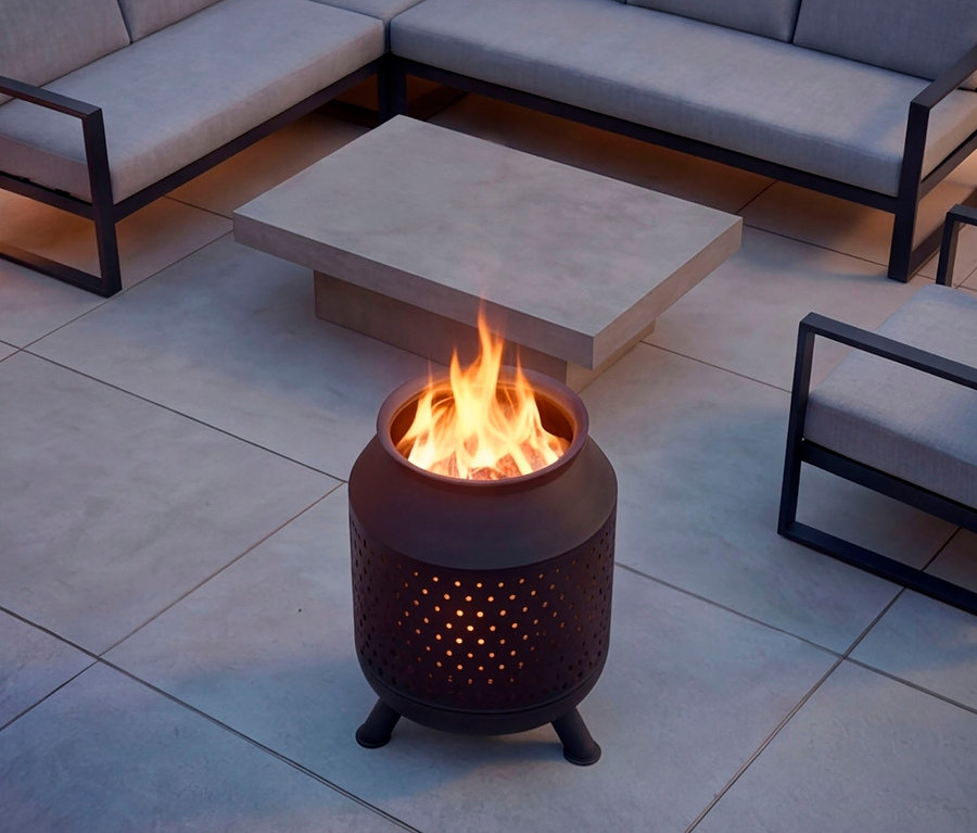Eine dunkle, zylindrische Feuerschale mit perforiertem Design und hellen Flammen steht auf einer gefliesten Terrasse, umgeben von einem hellgrauen Betontisch und einem Ecksofa mit hellen Polstern.