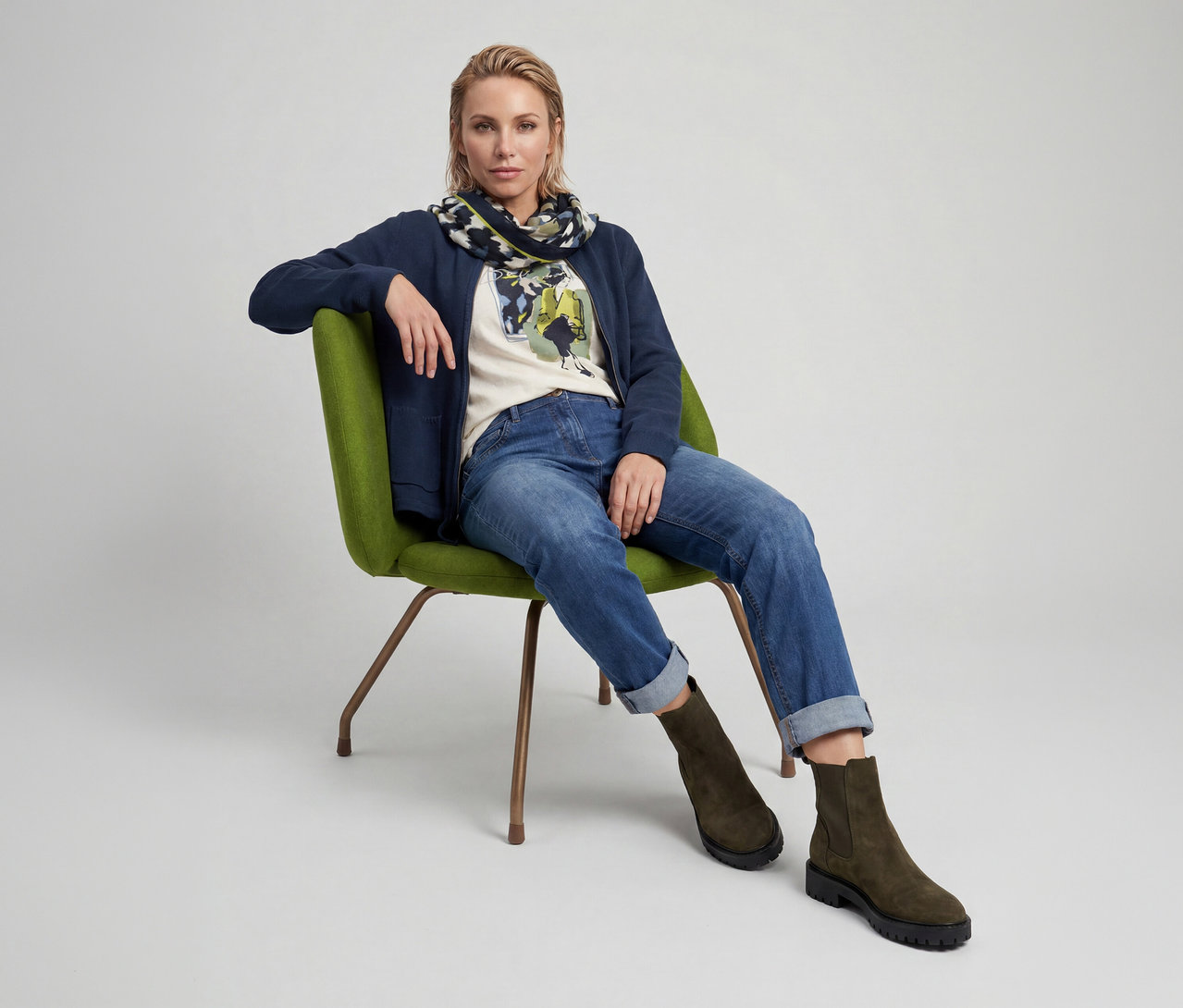 Eine blonde Frau sitzt in einem grünen Sessel und trägt eine marineblaue Jacke, ein cremefarbenes T-Shirt mit Aufdruck, hochgekrempelte blaue Jeans, einen gemusterten Schal und olivgrüne Stiefel.