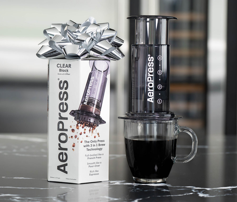 AeroPress Coffee Maker »Clear«, schwarz mit silberner Schleife auf einer Marmoroberfläche, daneben eine Tasse Kaffee.