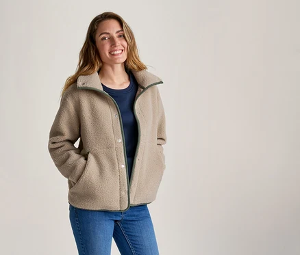 Lächelnde Frau in beiger Fleecejacke mit grünem Besatz, blauem T-Shirt und Jeans, die Hände in den Jackentaschen.