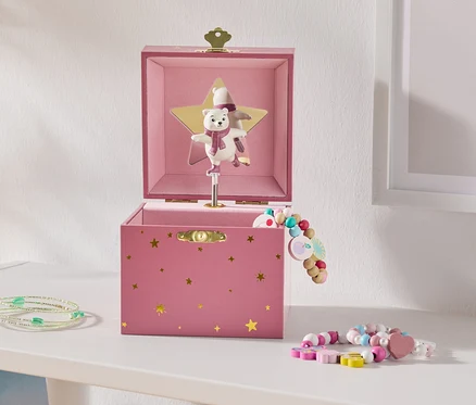 Eine offene rosa Spieluhr mit goldenen Sternen und einem tanzenden Eisbären auf einem goldenen Stern, daneben bunte Perlenarmbänder auf einem weißen Regal.