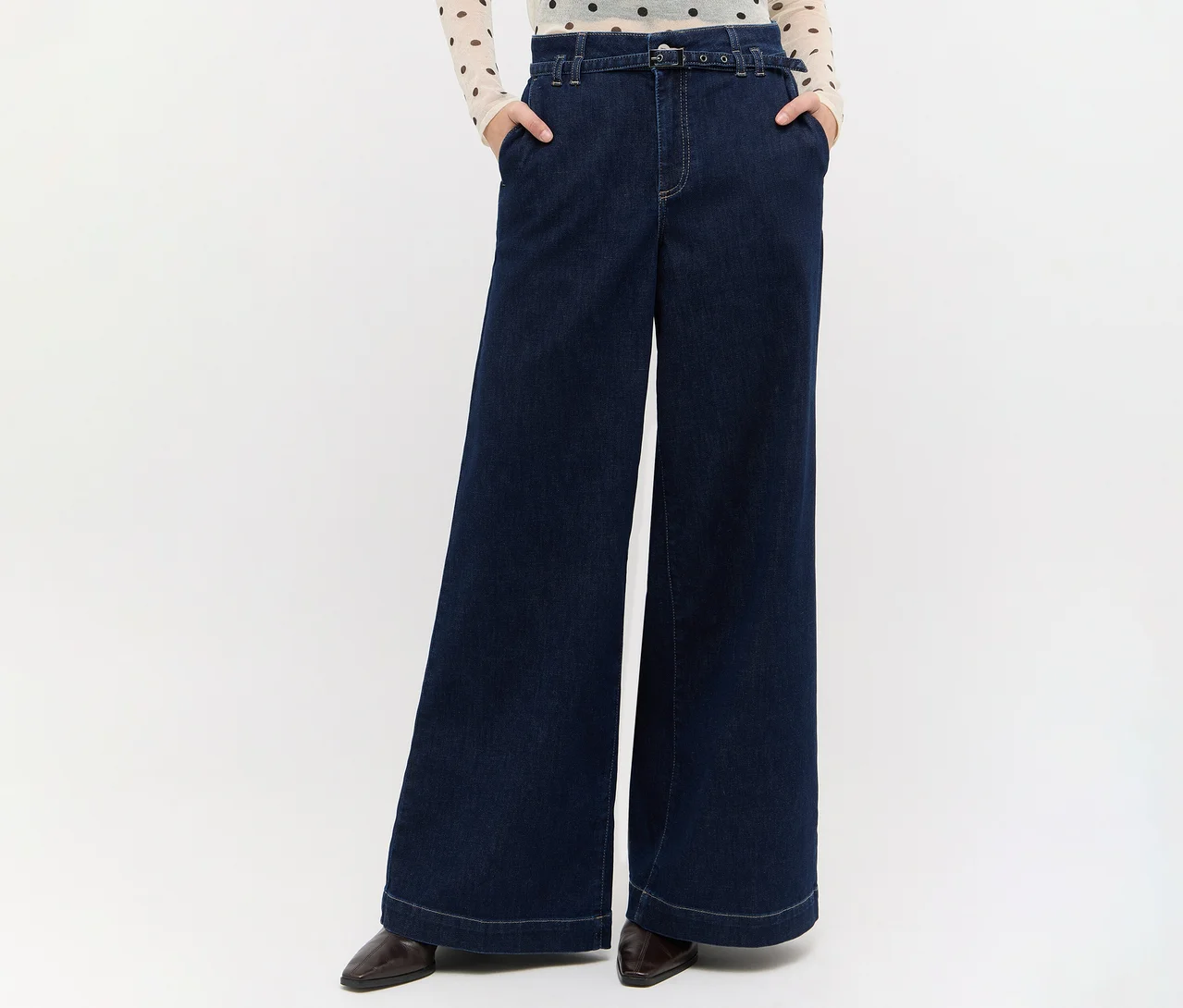 Person in dunkelblauer weiter Jeans mit Gürtel, hellem gepunktetem Oberteil und dunklen Schuhen, Hände in den Taschen.