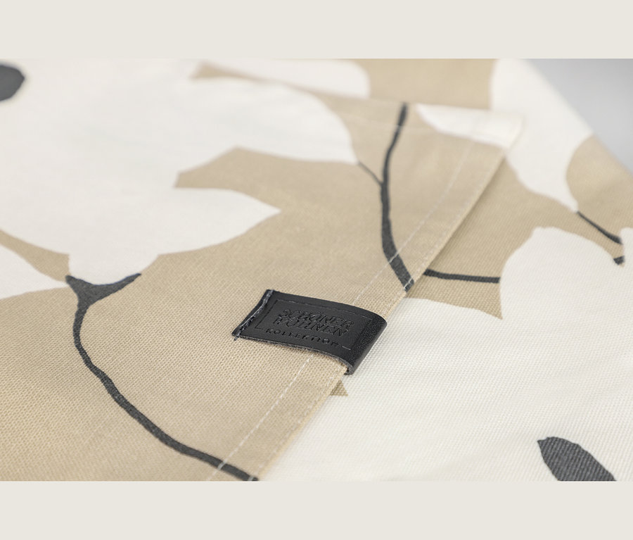 Nahaufnahme eines Stoffes mit floralem Muster in Beige, Weiß und Dunkelgrau, mit einem schwarzen Etikett mit der Aufschrift „SCHÖNER WOHNEN KOLLEKTION“.