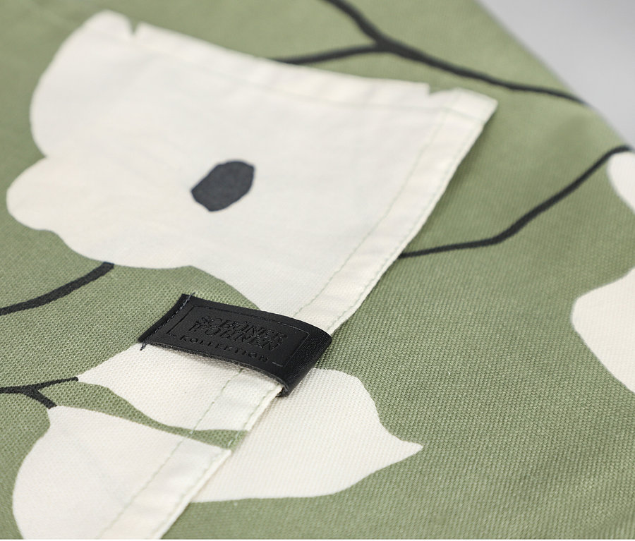 Nahaufnahme eines khakifarbenen SCHÖNER WOHNEN-Kollektion Tischläufers mit weißem Blumenmuster und schwarzen Umrissen. Ein schwarzes Etikett mit 'SCHÖNER WOHNEN KOLLEKTION' ist auf den Stoff genäht.
