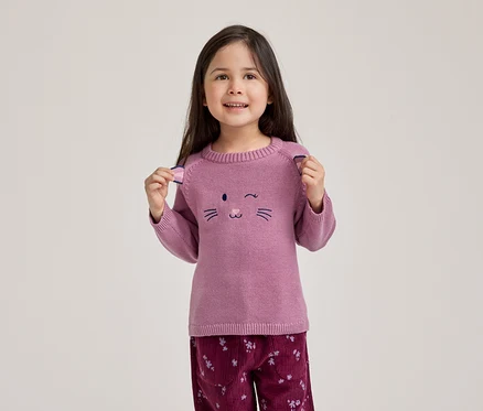 Ein lächelndes kleines Mädchen mit dunklen Haaren trägt einen rosa Pullover mit einem zwinkernden Katzengesicht und eine dunkelrote Cordhose mit Blumenmuster. Sie hält die kleinen Ohren-Details an den Schultern ihres Pullovers hoch.