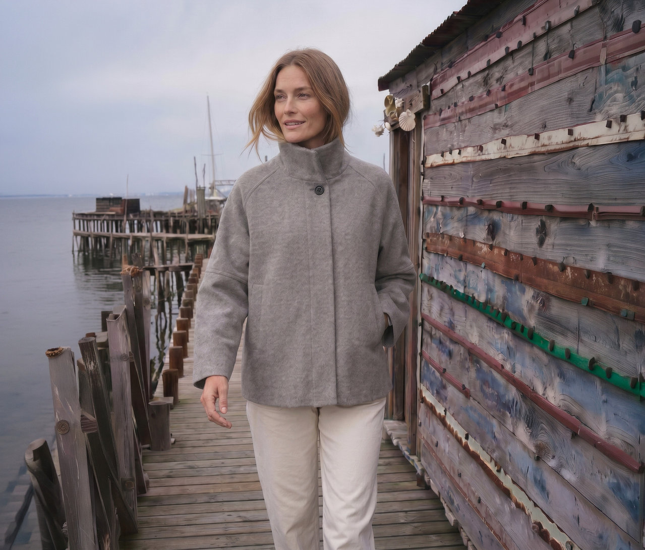 Frau geht auf einem Holzsteg, trägt eine graue Jacke mit Stehkragen und helle Hose, neben einer rustikalen Holzhütte mit Wasser und einem entfernten Steg im Hintergrund.
