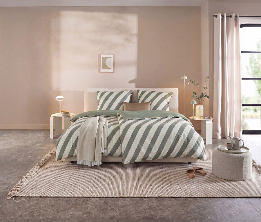 Ein Schlafzimmer mit einem Bett, das mit grün-weiß gestreifter SCHÖNER WOHNEN Renforcé-Bettwäsche bezogen ist. Auf dem Boden liegt ein heller Teppich, daneben ein Hocker mit Teekanne.