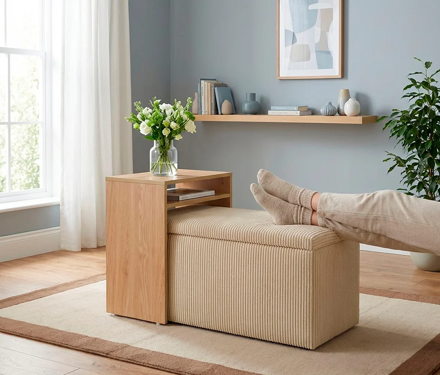Beine einer Person mit hellen Socken ruhen auf einem beigen, gerippten Ottomanen. Daneben steht ein Holztisch mit einer Vase weißer Blumen. Der Raum hat eine blaue Wand mit einem Regal, ein Fenster mit Vorhängen und eine Topfpflanze.