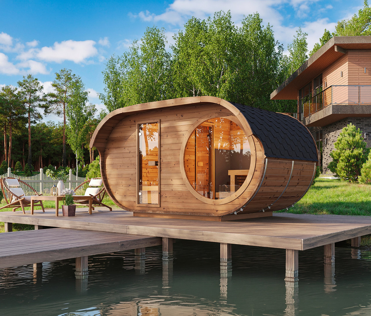 Hölzerne Fasssauna auf einer Holzterrasse über Wasser, mit Gartenmöbeln, umgeben von Bäumen und einem Haus im Hintergrund.