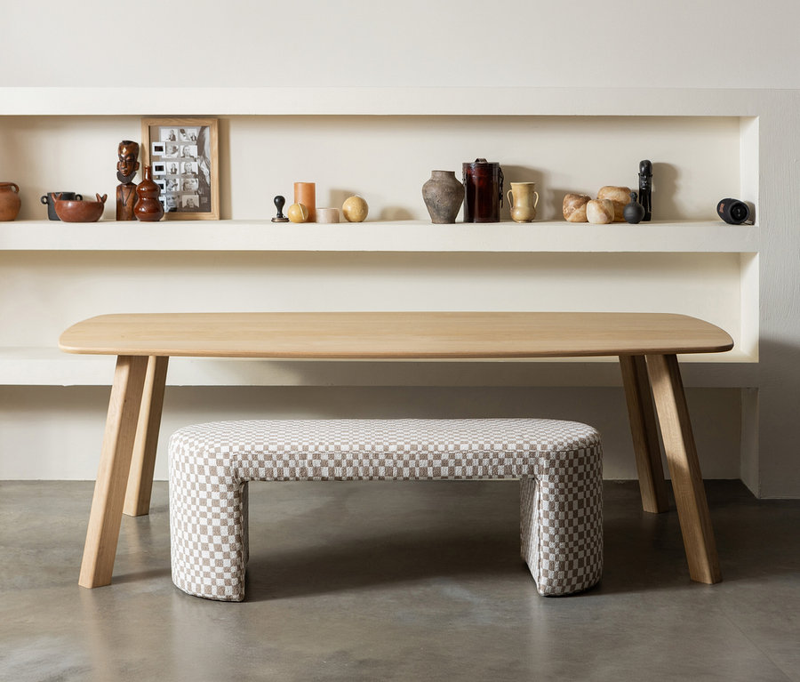 Ein naturfarbener WOOOD Tisch »Tablo« (ca. 180 x 100 cm) mit abgerundeter Platte und schrägen Beinen, darunter eine karierte U-förmige Bank. Auf den Regalen stehen verschiedene Dekoartikel.