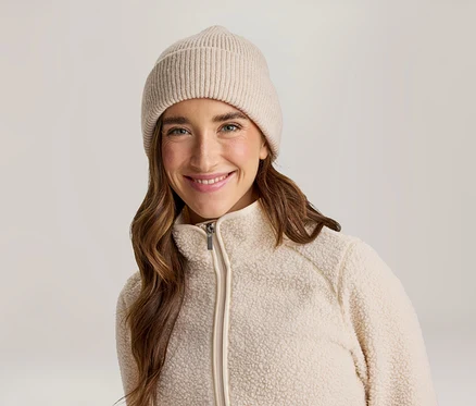 Lächelnde Frau mit langen braunen Haaren, die eine beige gerippte Mütze und eine cremefarbene Fleecejacke mit Reißverschluss trägt.