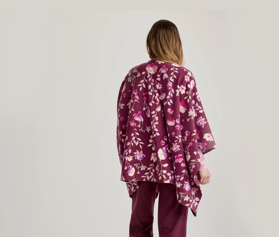 Eine Person von hinten, die einen burgunderfarbenen floralen Poncho mit rosa, lila und weißen Blumen und passende burgunderfarbene Hosen vor einem hellen Hintergrund trägt.