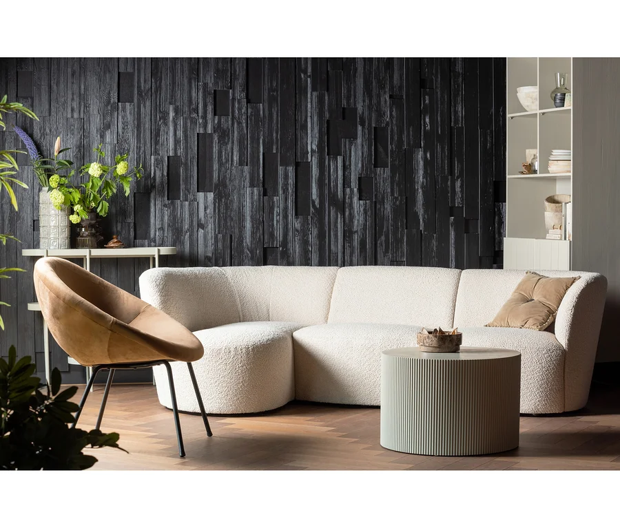 Ein modernes Wohnzimmer mit einem cremefarbenen L-förmigen Sofa, einem runden beigefarbenen Sessel und einem geriffelten hellgrünen Couchtisch auf einem Fischgrätparkett. Eine dunkle strukturierte Holzwand ist im Hintergrund, rechts steht ein weißes Regal.