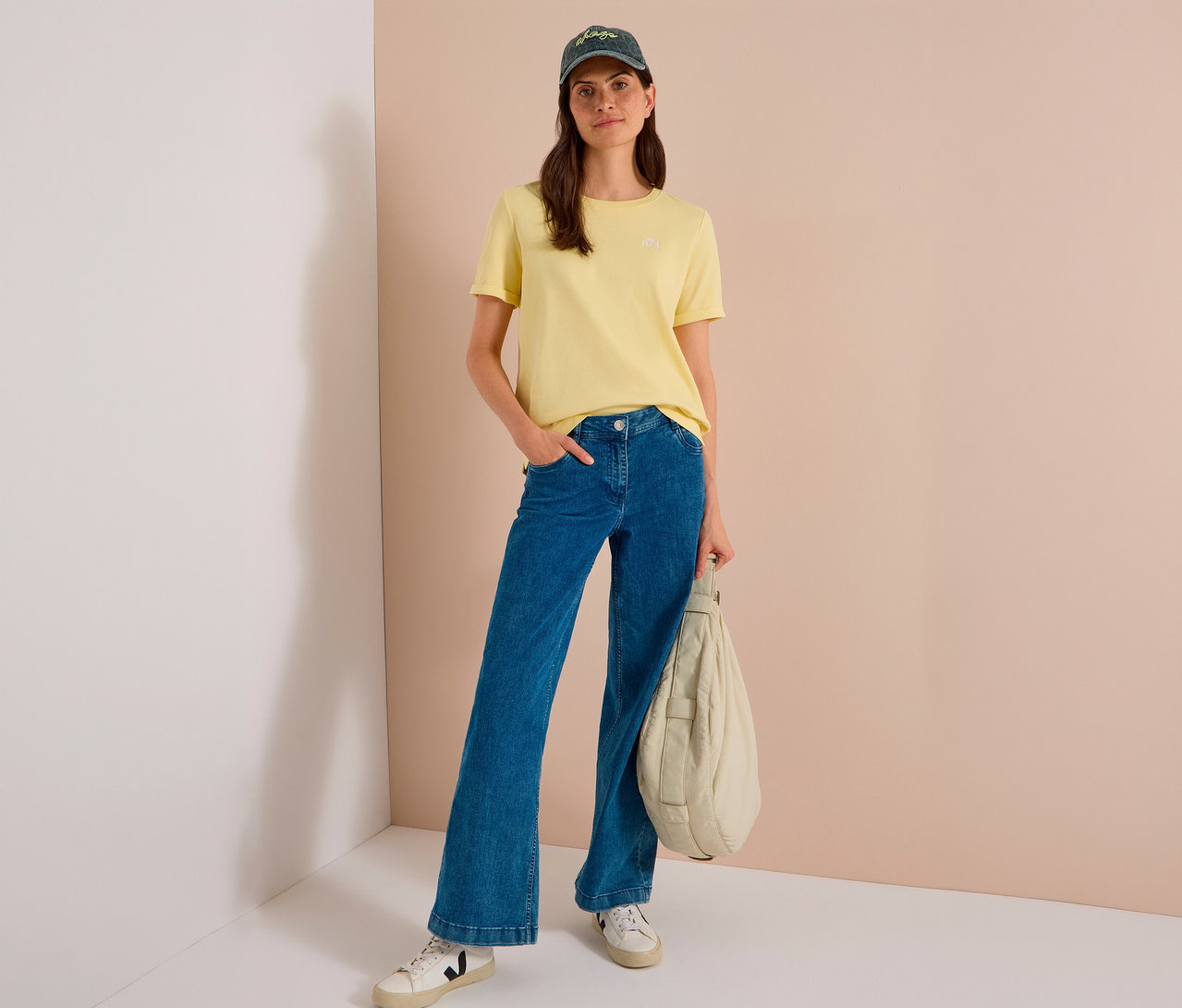 Frau trägt gelbes T-Shirt, blaue weite Jeans, weiße Sneaker und eine dunkelgrüne CECIL Baseballcap, hält eine hellfarbene, gepolsterte Tasche.
