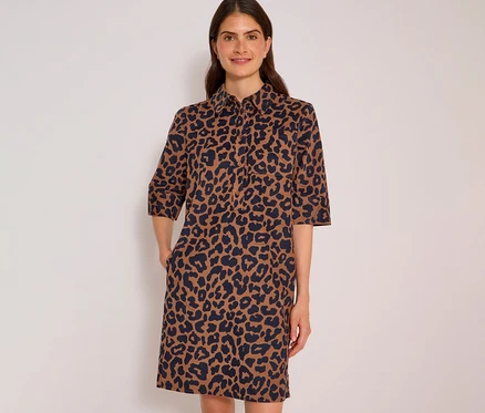 Lächelnde Frau in einem braun-dunkelblauen CECIL Hemdblusenkleid mit Leopardenmuster und Dreiviertelärmeln, Hände in den Taschen.