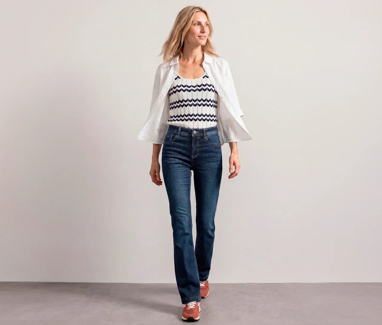 Eine blonde Frau geht in einer weißen Bluse über einem weiß-marineblauen Tanktop und Street One Bootcut Jeans.