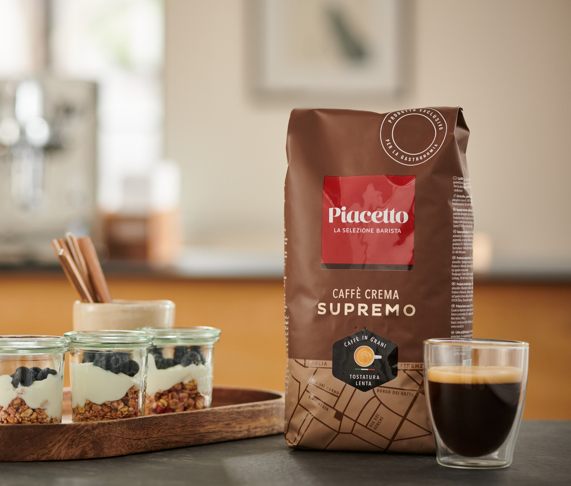 Auf einem Tisch stehen Müsligläser mit Blaubeeren, eine Packung Piacetto Supremo Caffè Crema und eine Tasse Espresso.