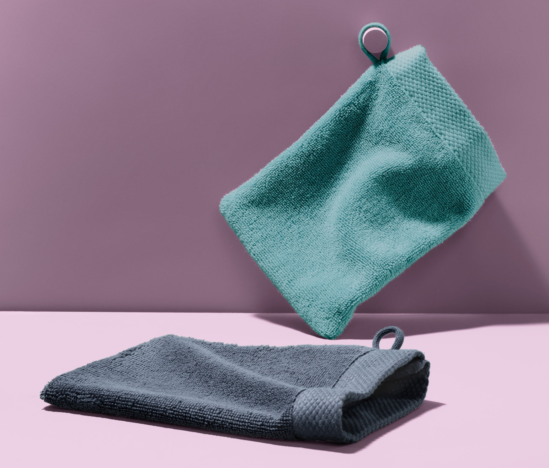 Zwei Waschlappen vor einem lila Hintergrund. Ein hellblauer Waschlappen hängt in der Luft und ein dunkelgrauer Waschlappen liegt auf der Oberfläche.
