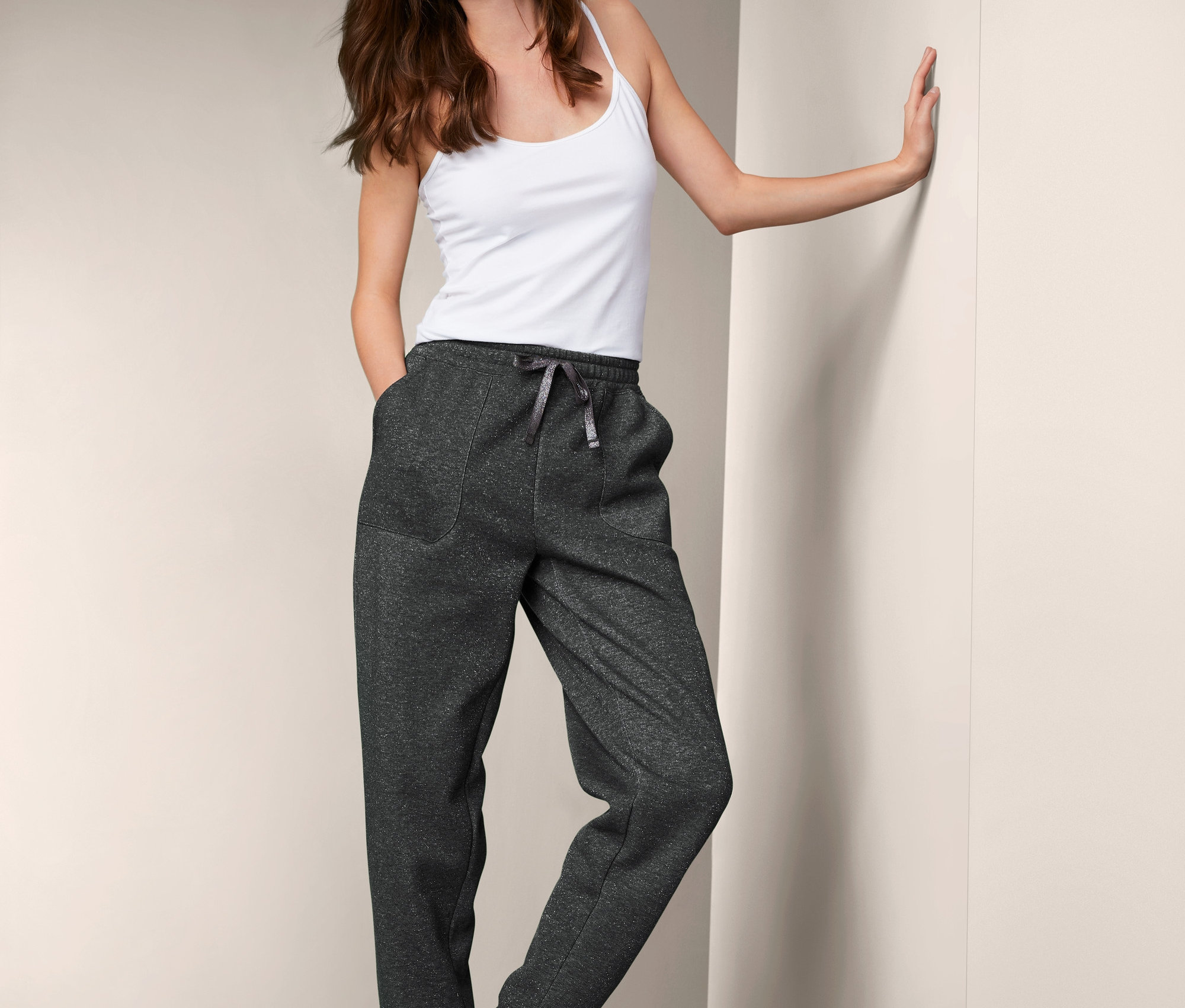 Lächelnde Frau lehnt an der Wand und trägt ein Tanktop und eine Loungewear-Hose.