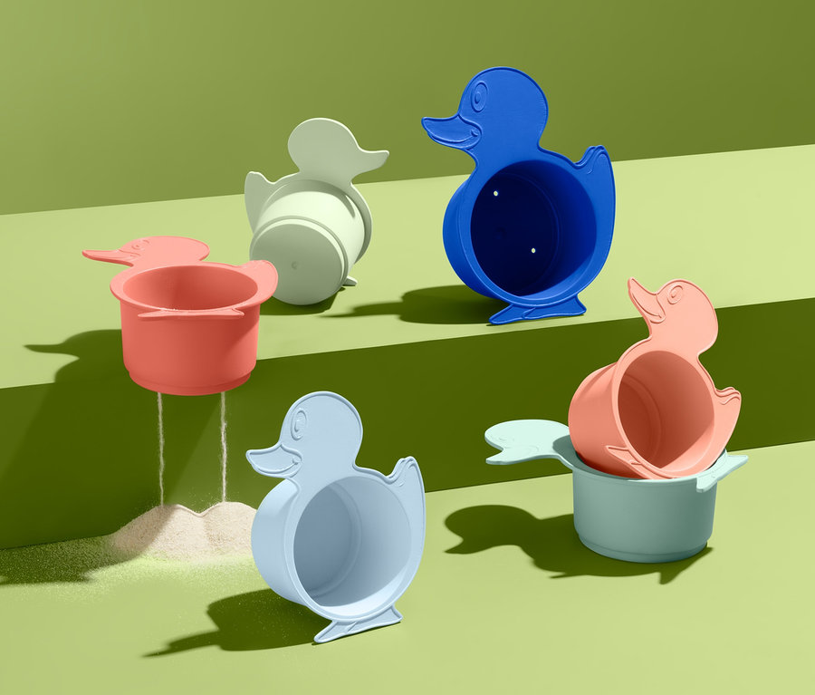 Sechs bunte Bade- und Sandspielbecher in Entenform stehen auf grünem Grund, aus einem rieselt Sand.